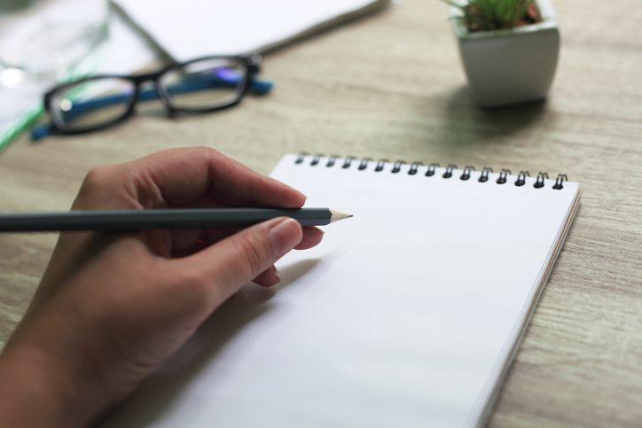 Female hand with pencil writing in the notepad on desk. Natural daylight. International Lefthanders Day.