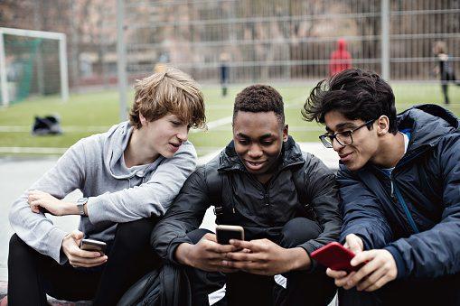 Smiling friends looking at teenage boy’s phone while sitting against soccer field